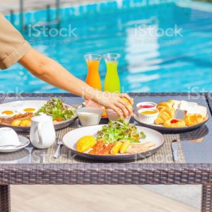 Waiter hand serving breakfast in restaurant at luxury hotel. Asian woman waitress preparing morning food and drinks on table in outdoors pool cafe in tropical resort. Travel, holiday, vacation concept
