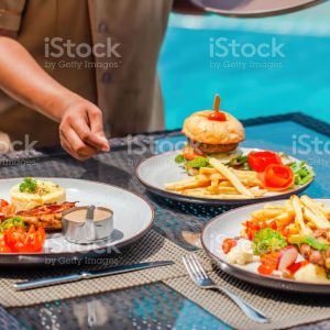 Waitress hand serving lunch in restaurant at luxury hotel. Asian woman waiter put delicious food plates on table in outdoors pool cafe in tropical resort. Summer travel, holidays, vacations concept.